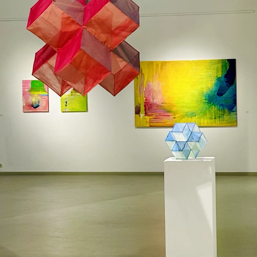 Drei Gemälde von Joachim Kersten im Hintergrund der Ausstellung „Augenlust“, davor schwebende geometrische Skulpturen.